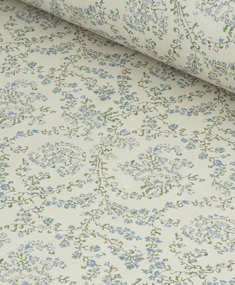 A close-up of a roll of wallpaper with a delicate floral pattern in light blue and green on a cream background. The design features tiny flowers and vines arranged in swirling, intricate shapes.
