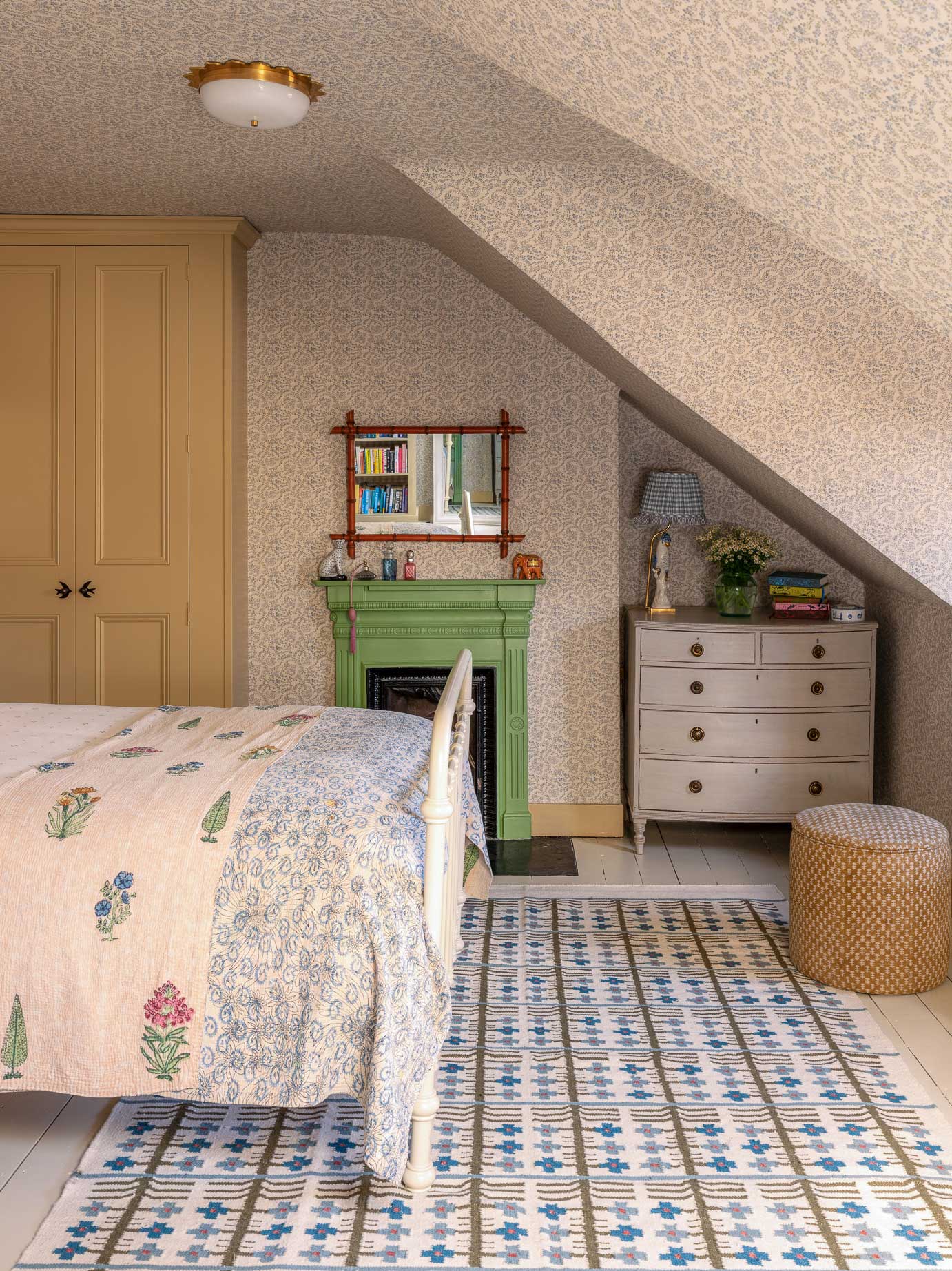 Cozy attic bedroom with a sloped ceiling, patterned rug, bed with floral quilt, green fireplace, wall mirror, cream dresser with lamp and flowers, and a woven ottoman. Warm, neutral tones create a welcoming atmosphere.