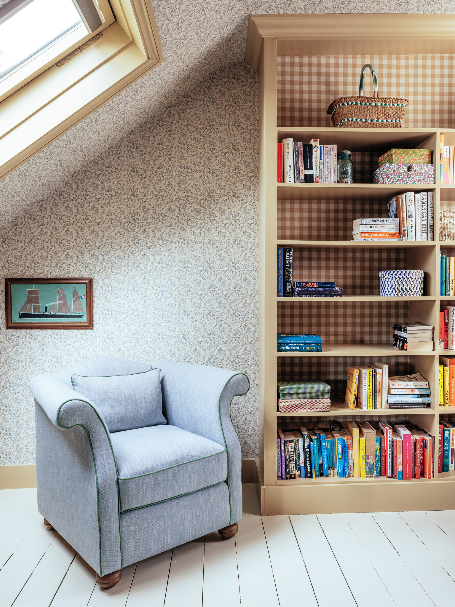 A cozy reading nook with a light blue armchair next to a tall built-in bookshelf filled with colorful books, under a slanted ceiling with a skylight window. A small framed picture hangs on the wall.