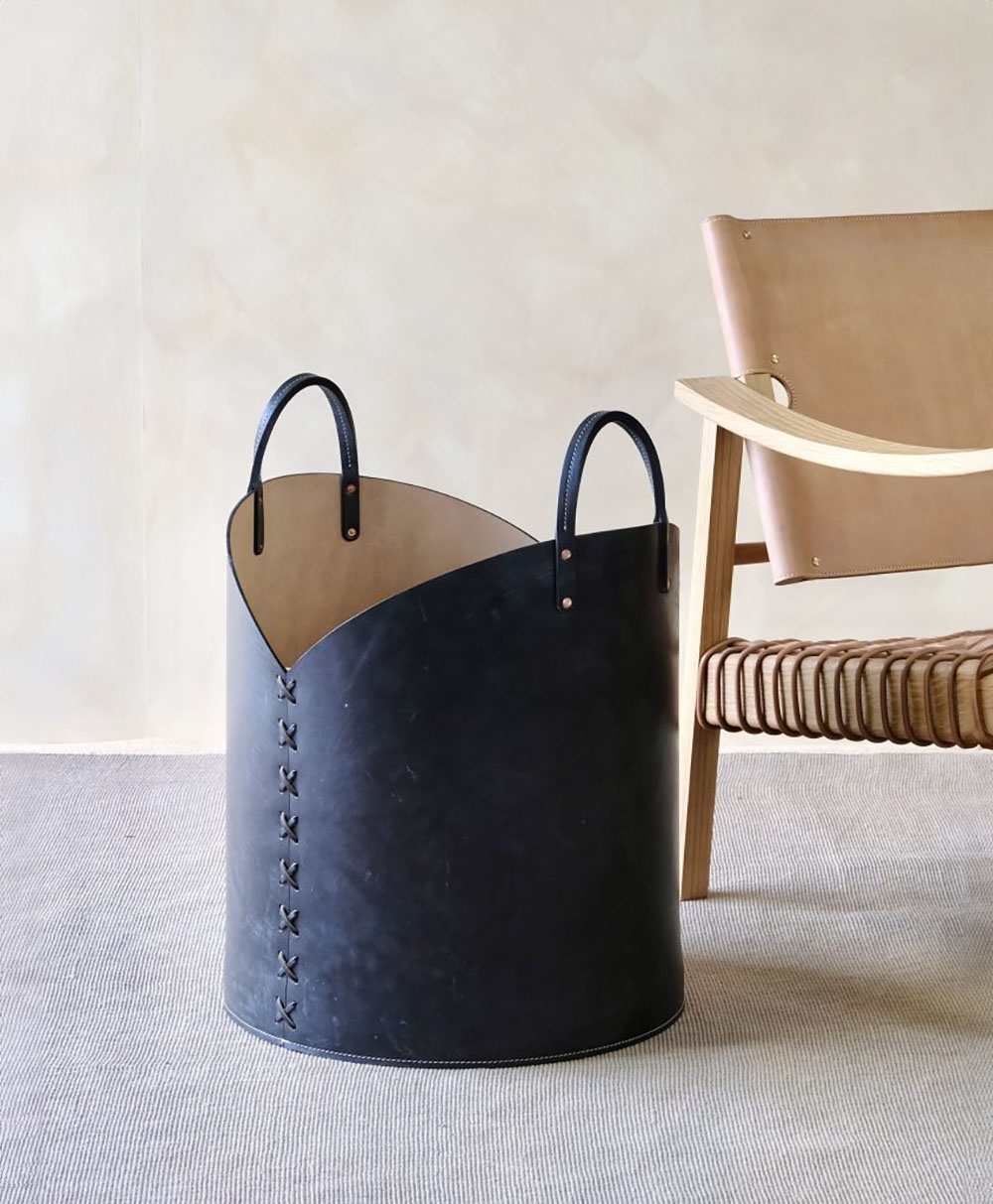 A modern black leather storage basket with handles sits on a light carpet next to a woven, light wood and tan leather chair against a neutral wall.