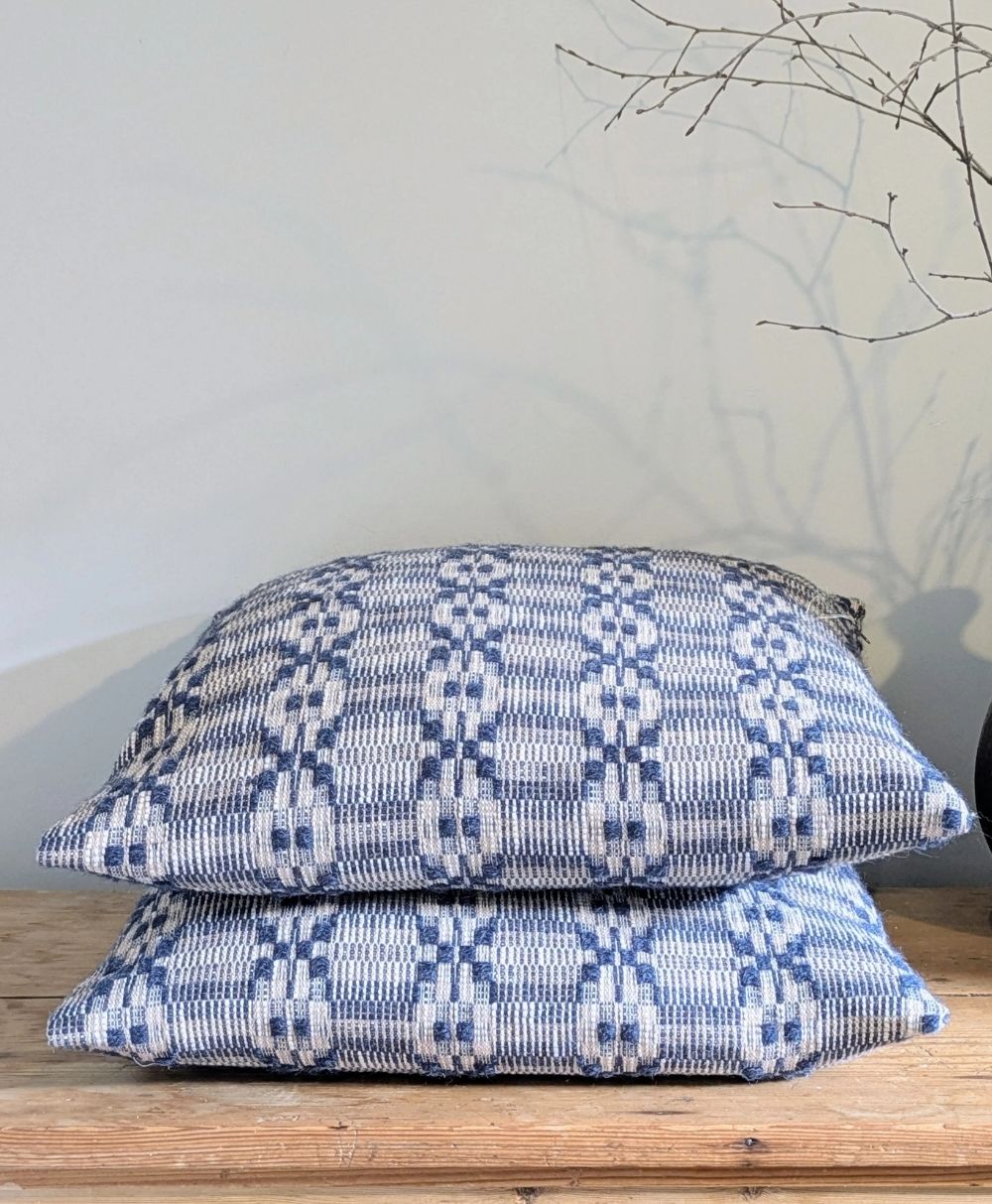 Two blue and white patterned cushions stacked on a wooden surface, with a minimalist background and a branch in a black vase partially visible on the right.