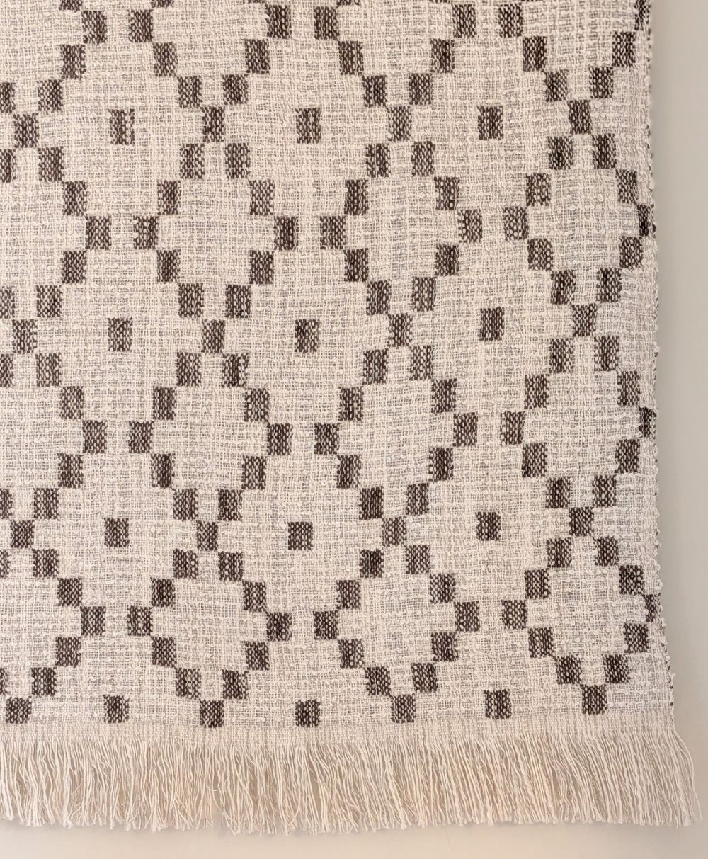 A close-up of a woven rug with a beige background and a repeating geometric diamond pattern in brown. The rug has a fringe along the bottom edge.