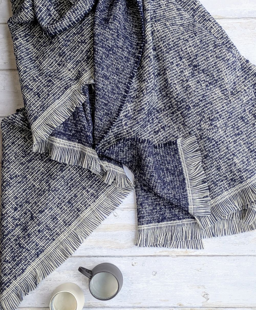 A textured blue and white fringed blanket is spread on a white wooden surface, with a gray mug and a white mug placed nearby, both containing drinks.