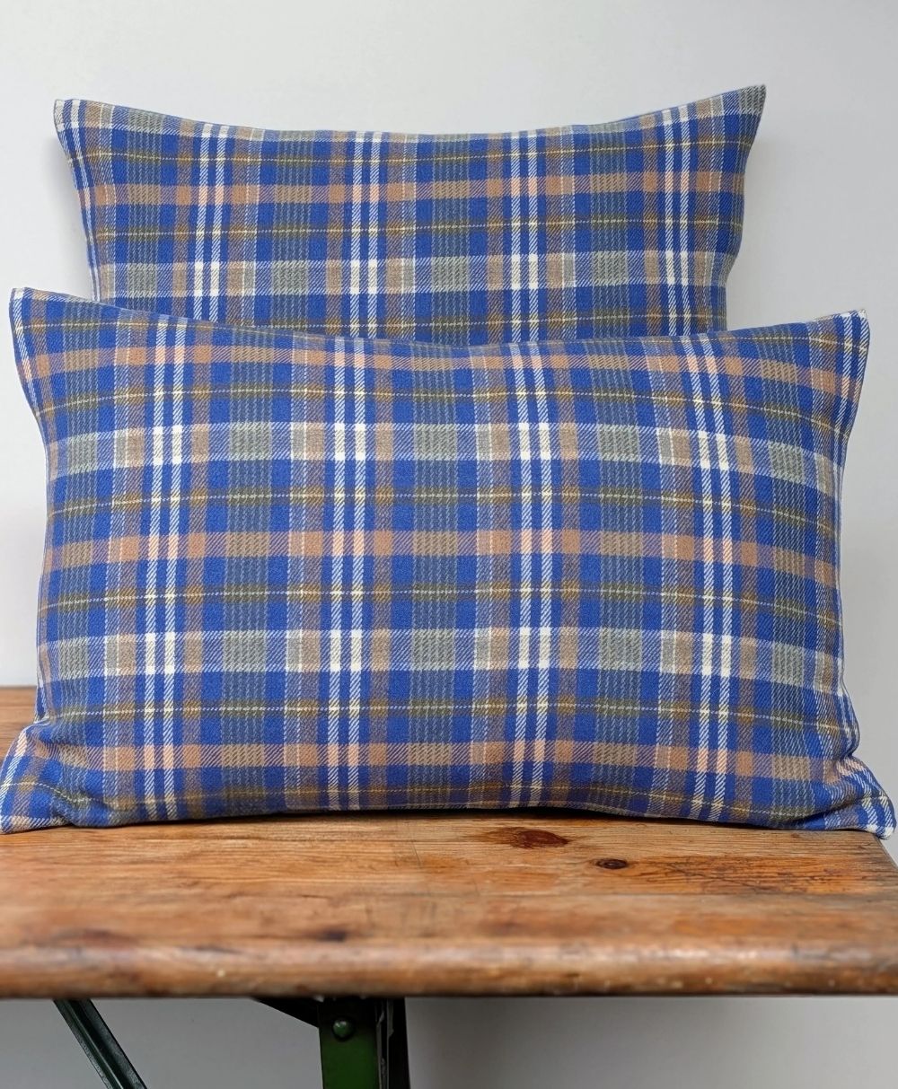 Two rectangular pillows with a blue, brown, and white plaid pattern are stacked against a plain white wall, resting on a rustic wooden surface.