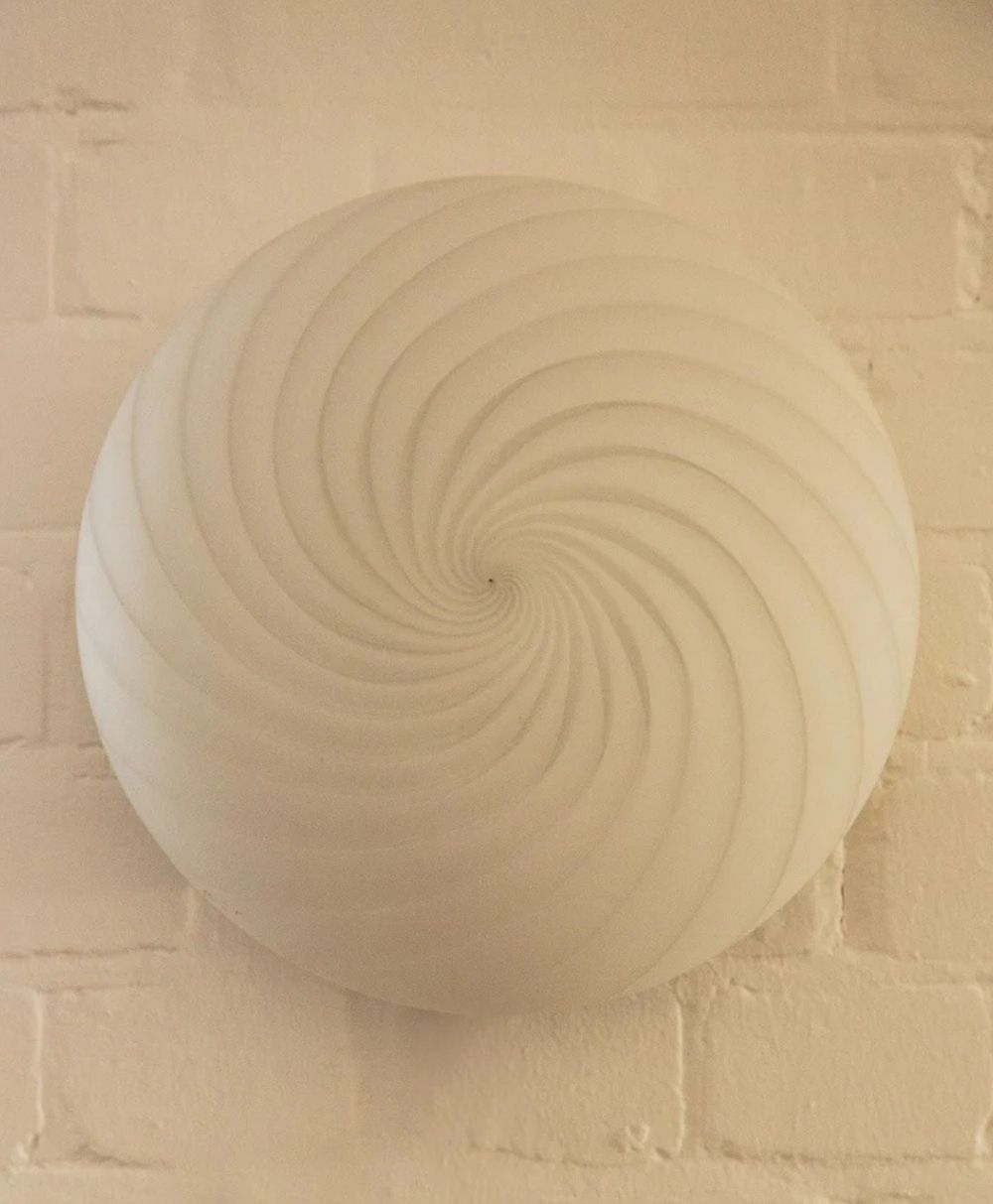 A round, cream-colored wall fixture with a spiral, shell-like pattern is mounted on a pale brick wall. The design features smooth, curved ridges that radiate from the center.