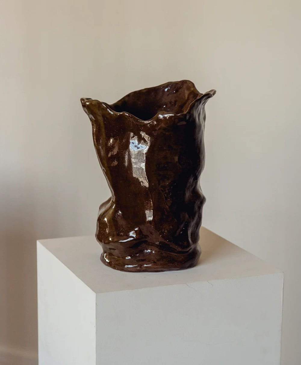 A glossy, dark brown ceramic vase with an irregular, wavy rim and textured surface sits on a white pedestal against a plain beige background.