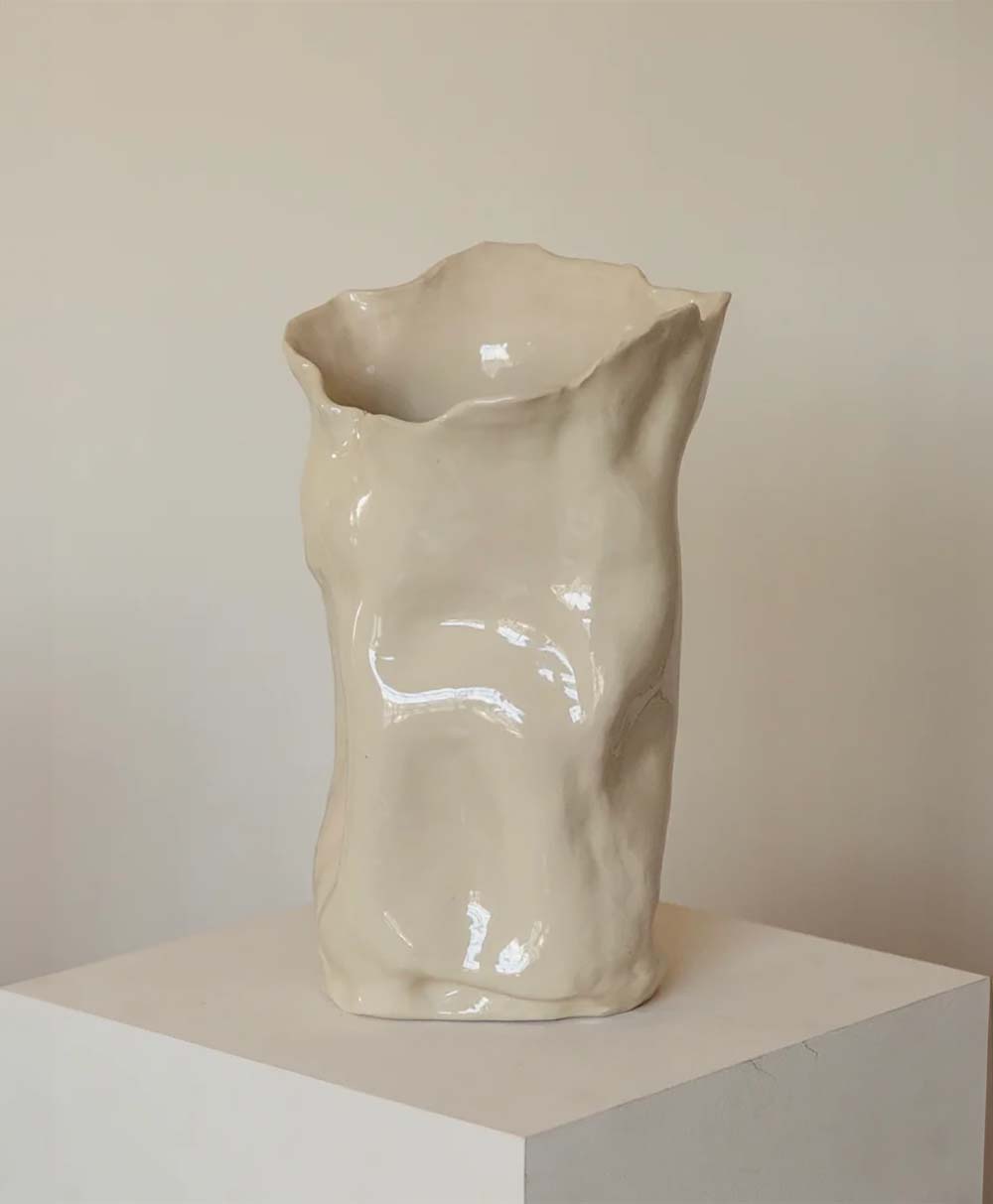 A glossy, off-white ceramic vase with an irregular, crumpled shape sits on a white pedestal against a plain beige background.