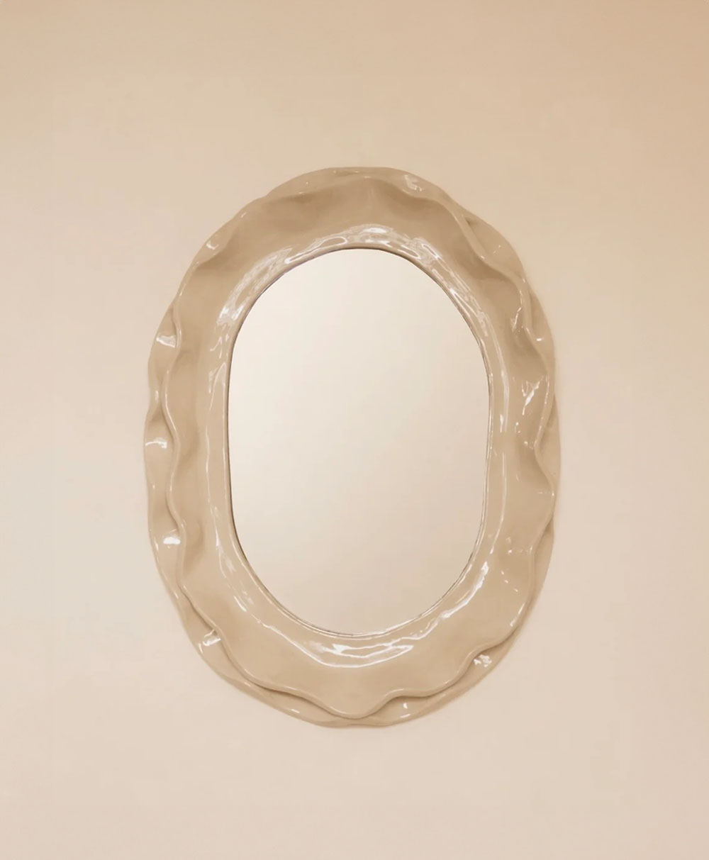 An oval mirror with a wavy, textured beige frame is mounted on a plain light-colored wall.
