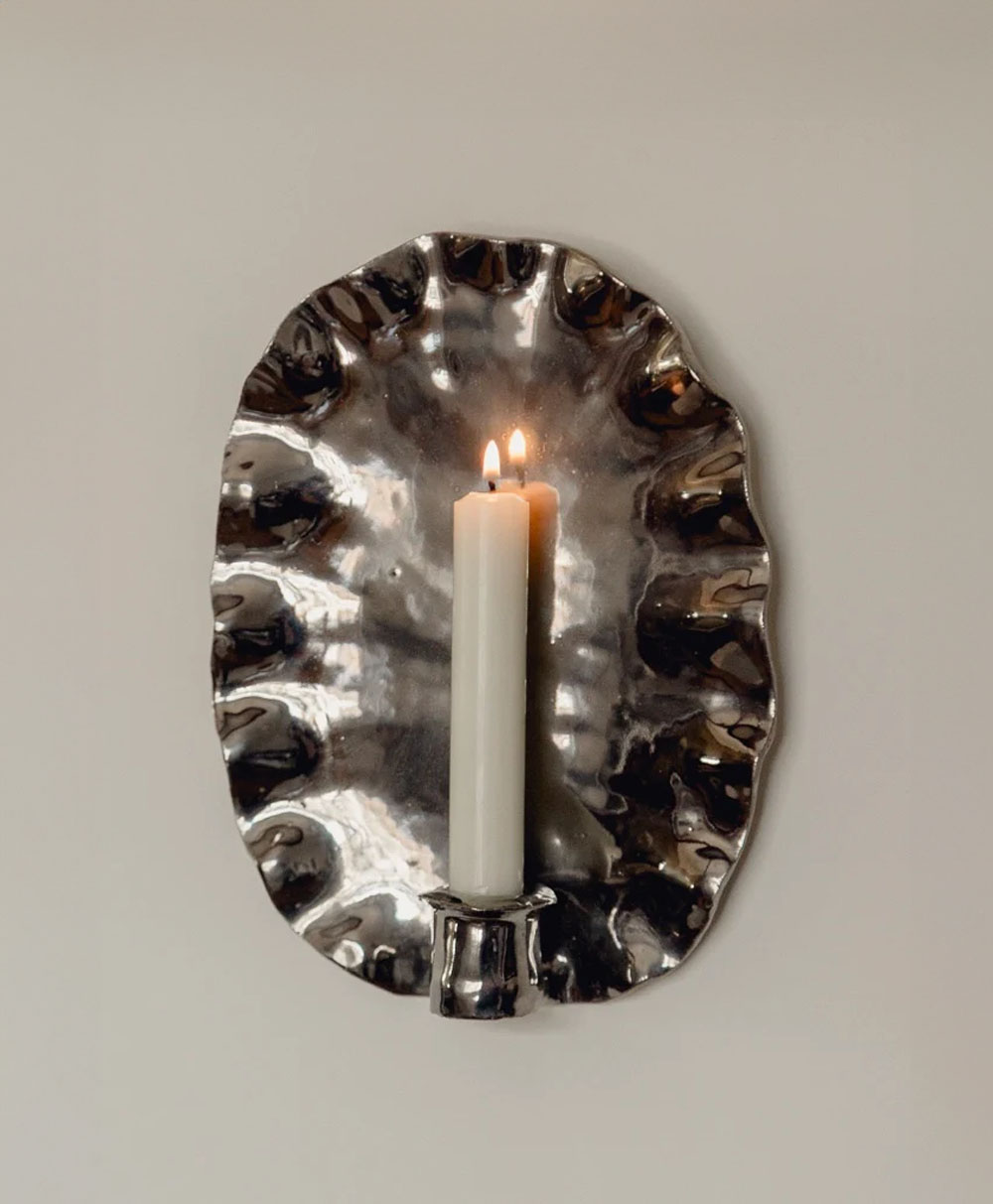 A white candle burns with two flames on a shiny, irregularly shaped metallic wall sconce mounted against a plain light-colored wall.