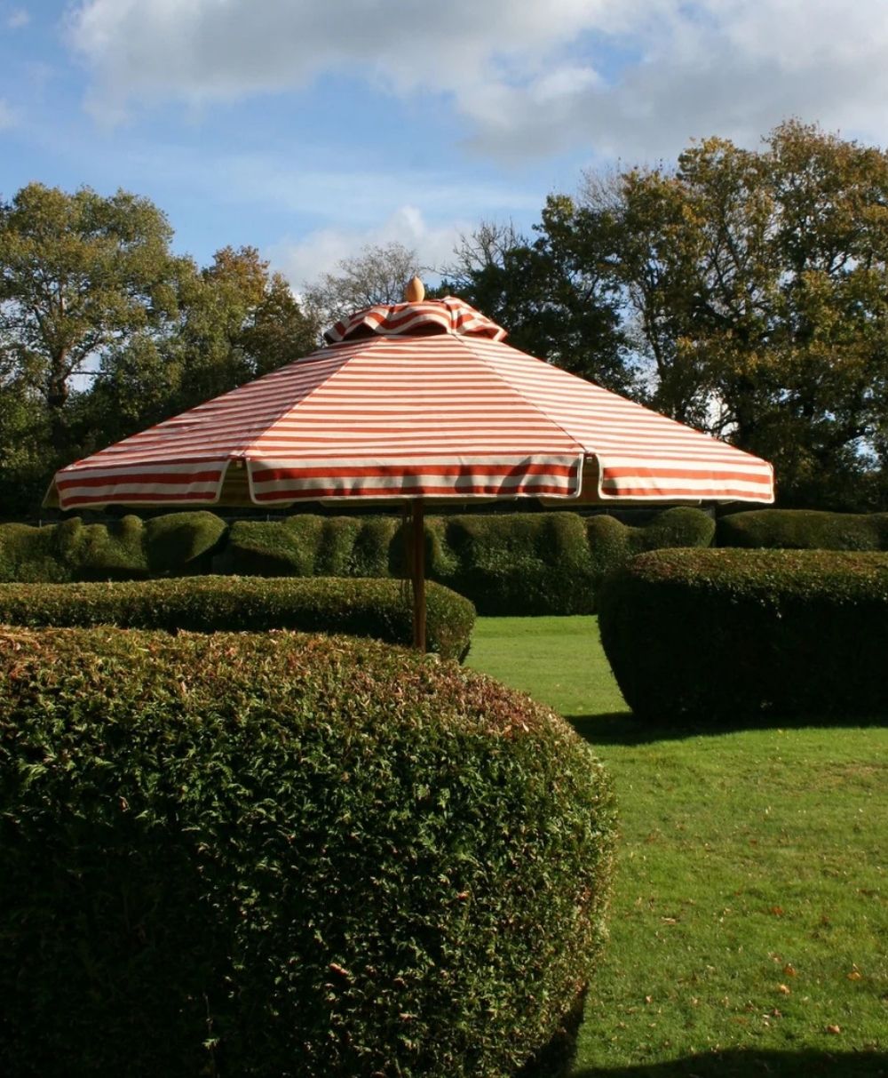 A red and white striped patio umbrella is set up in a garden with neatly trimmed hedges and green grass under a partly cloudy sky.