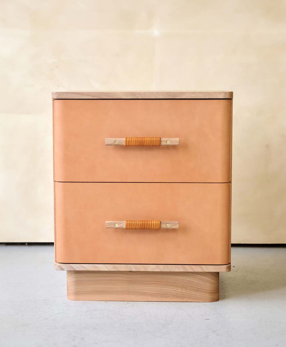 A modern wooden nightstand with two tan leather-fronted drawers, each featuring a handle wrapped with tan cord, set against a plain beige background.