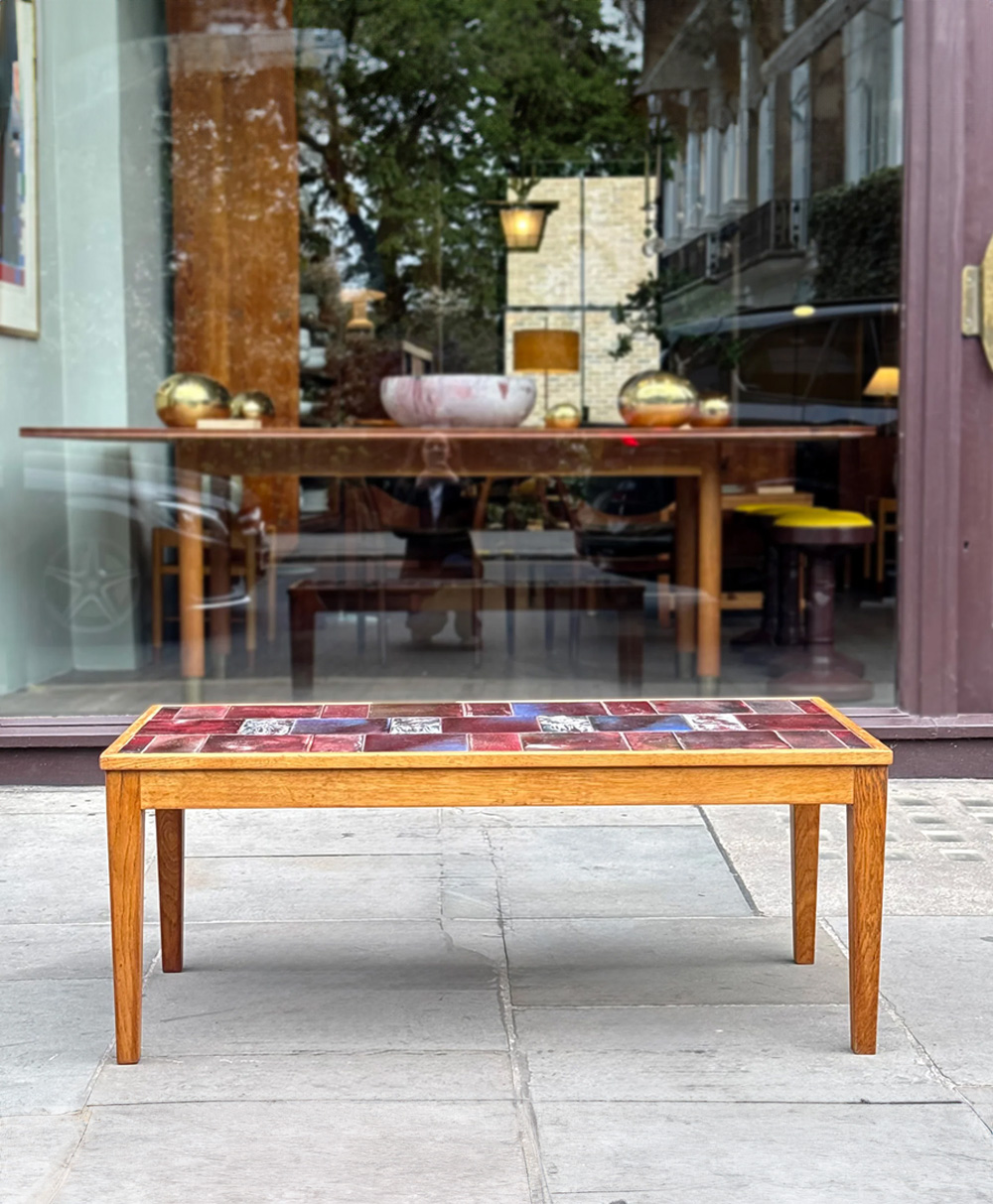 A rectangular wooden coffee table with a colorful tiled top stands outside on a stone pavement in front of a glass window reflecting another table and chairs inside. Trees and buildings are visible in the background.