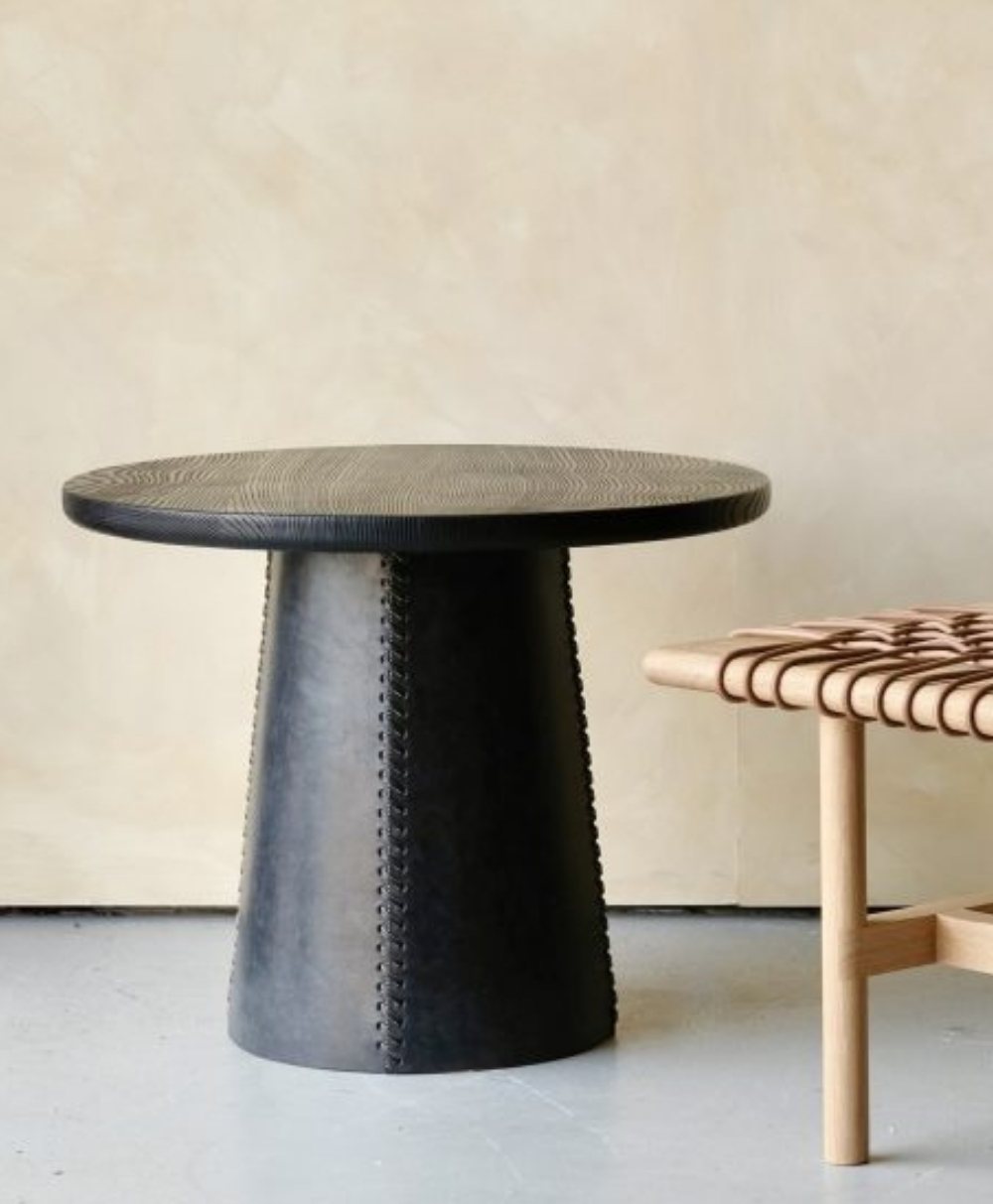 A round, dark wooden side table with a conical, textured base stands next to a light wooden bench with a woven leather seat, set against a neutral beige wall.