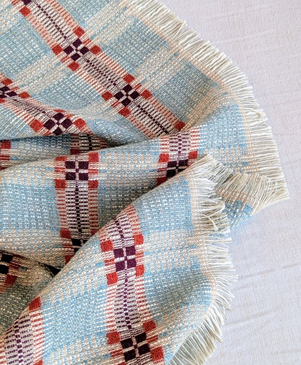 A close-up of a woven fabric with a plaid pattern in light blue, cream, red, and purple tones. The fabric has fringed edges and is laid out on a light-colored surface.
