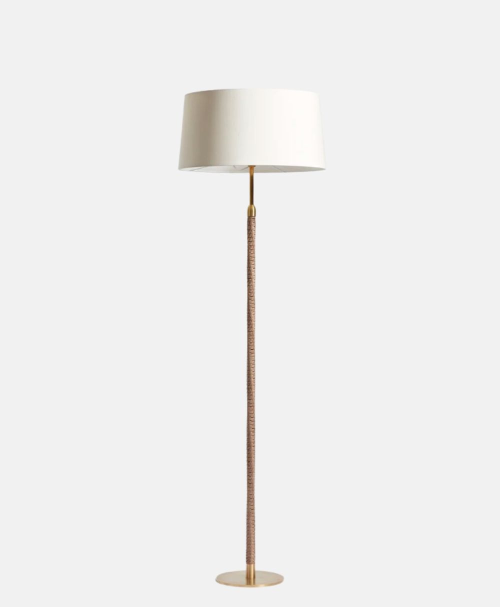 A modern floor lamp with a white drum lampshade, a slender pole wrapped in a textured brown material, and a round gold base, set against a plain light gray background.