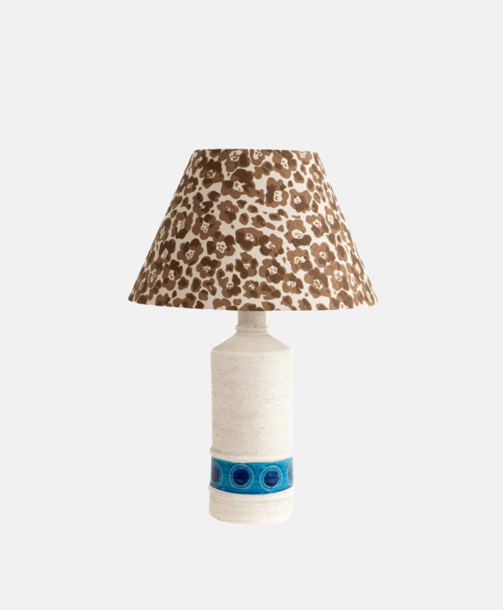 A table lamp with a textured off-white ceramic base featuring a blue circular pattern near the bottom and a wide, brown and white floral-patterned lampshade.