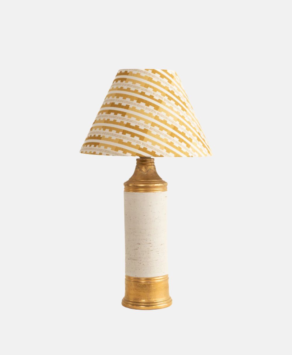 A table lamp with a cylindrical cream base featuring gold accents and a conical lampshade decorated with a yellow and white geometric pattern, set against a plain white background.