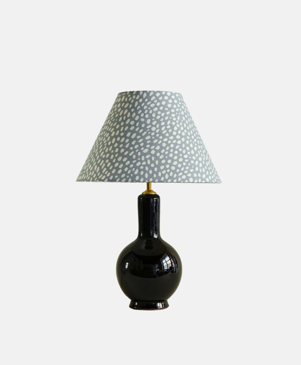 A table lamp with a glossy black, rounded base and a wide lampshade featuring a light blue color with a white, irregular dotted pattern. The lamp is set against a plain white background.