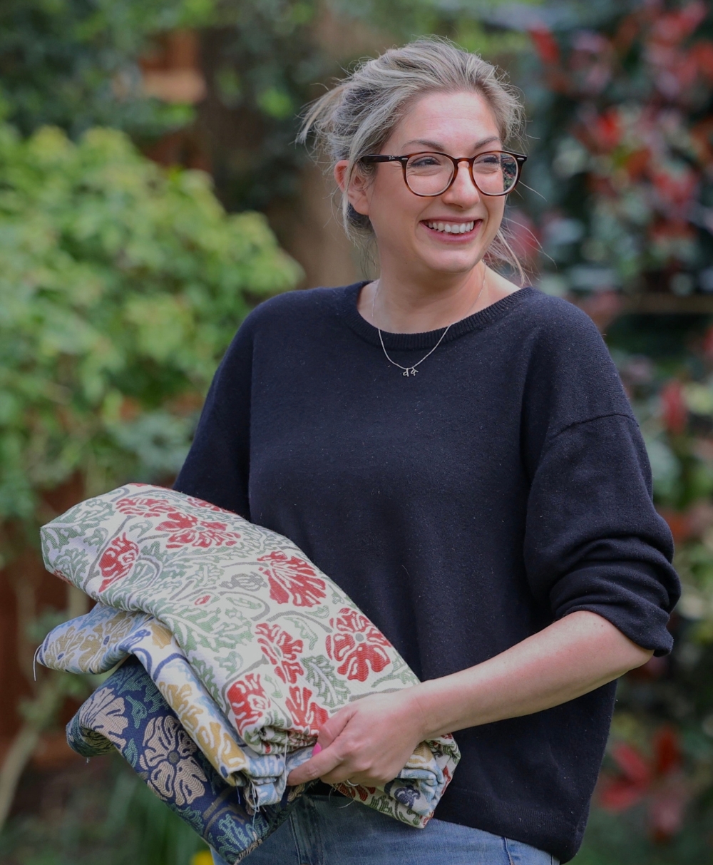 A smiling woman with glasses and a dark sweater stands outdoors, holding a stack of colorful, patterned fabrics with a blurred garden in the background.