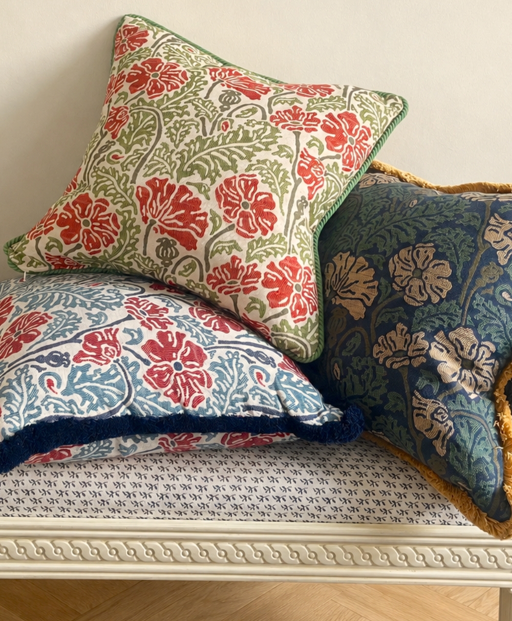 Three decorative pillows with floral patterns in red, green, and blue rest on a white cushioned bench with a subtle geometric design. The pillows have different colored backgrounds and borders.