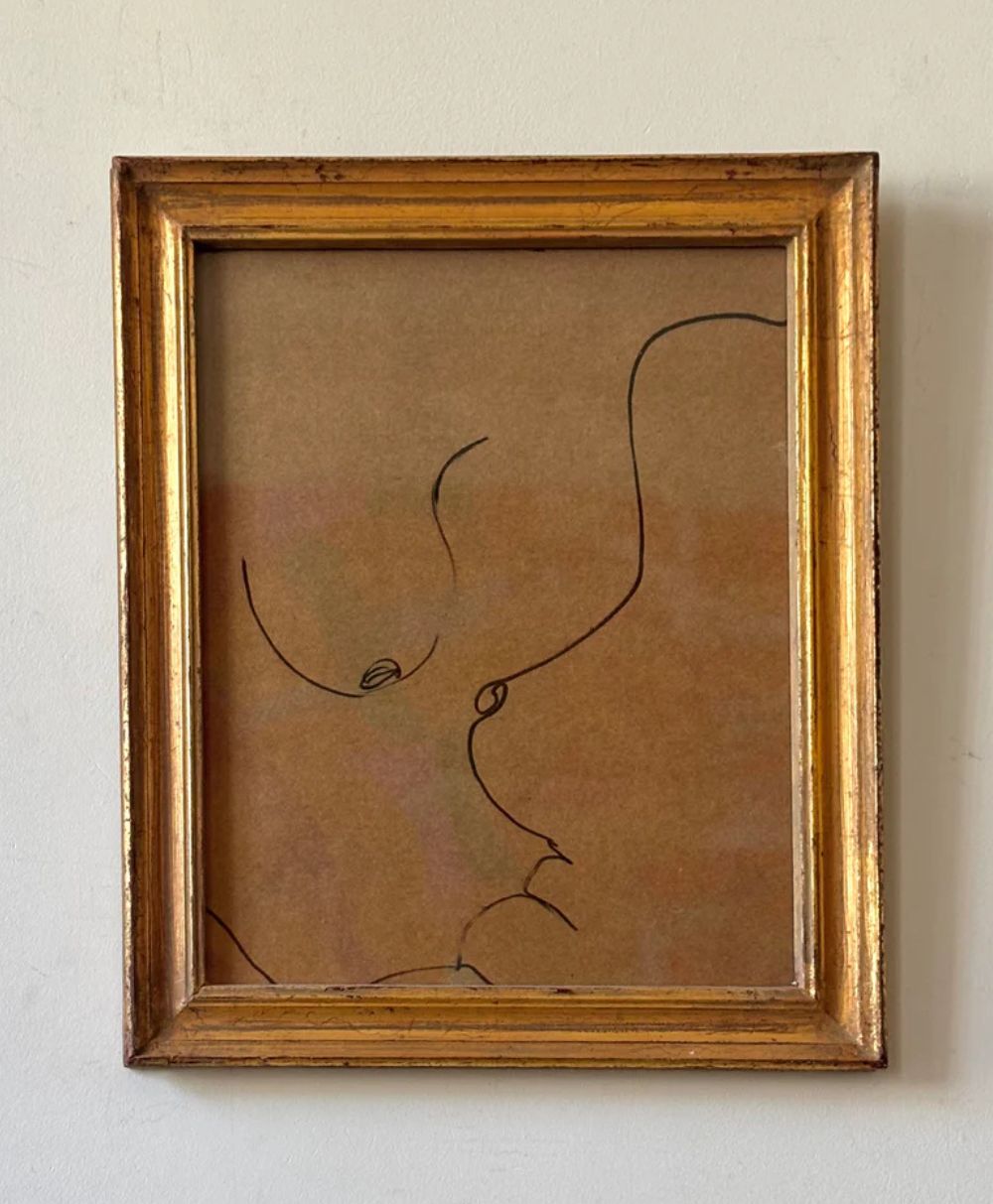 A minimalist line drawing of a nude figure on brown paper, framed in a simple gold frame, hangs on a white wall. The artwork uses a few elegant, continuous black lines.