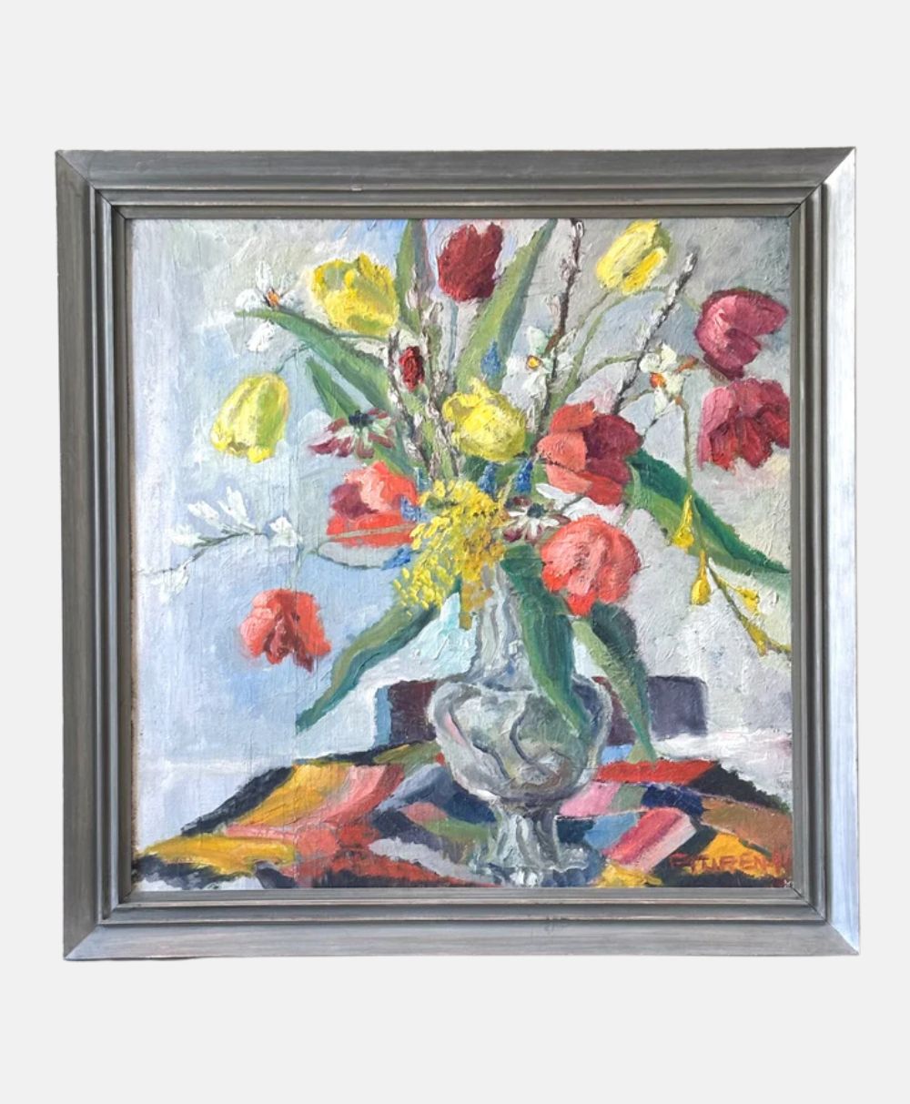 A framed painting of a glass vase filled with red and yellow flowers, set on a colorful patterned surface, with a light-colored background. The painting style is impressionistic with visible brushstrokes.