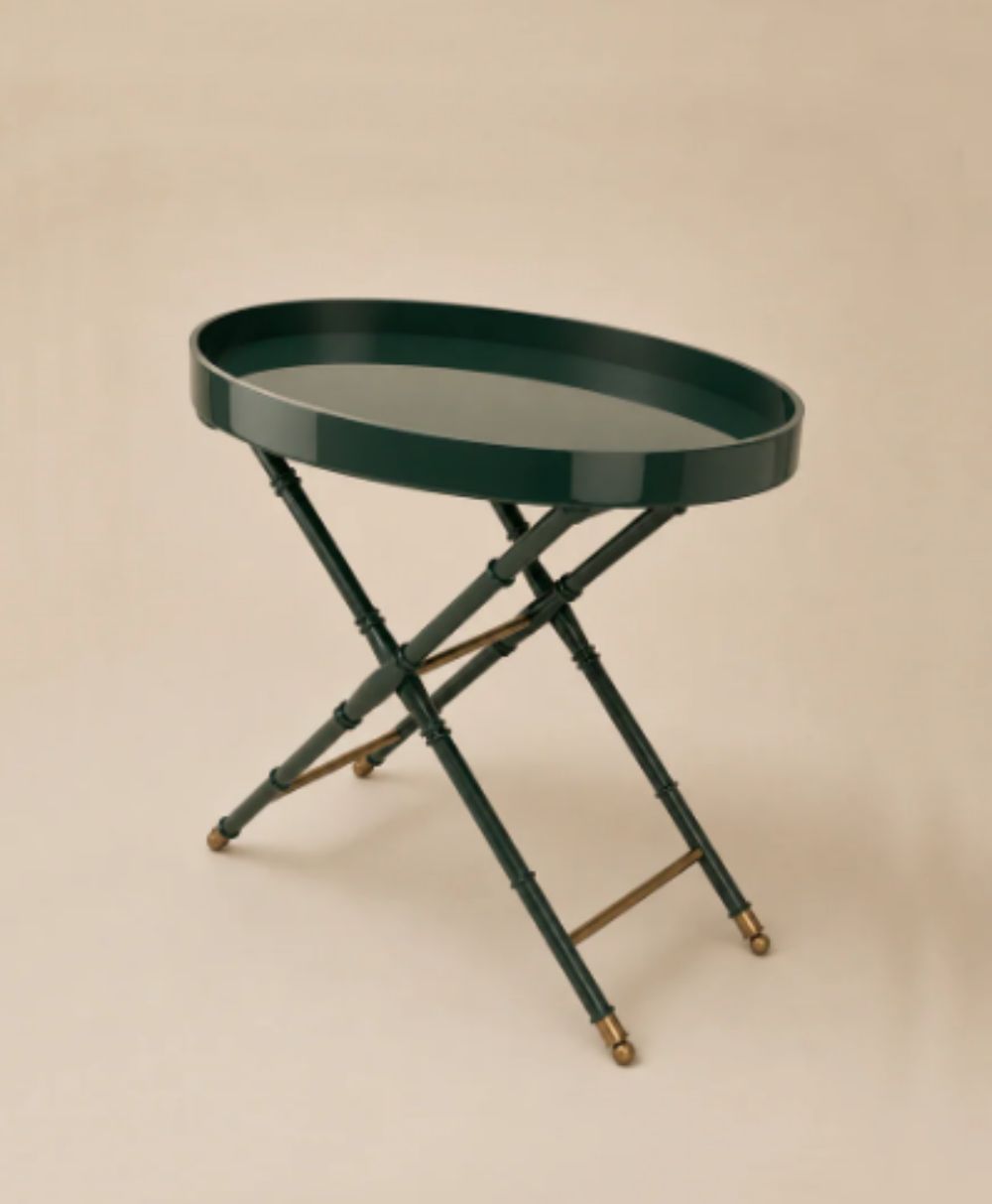 A dark green, round tray table with a raised edge, standing on crossed, spindle-style legs with gold accents at the feet, set against a beige background.