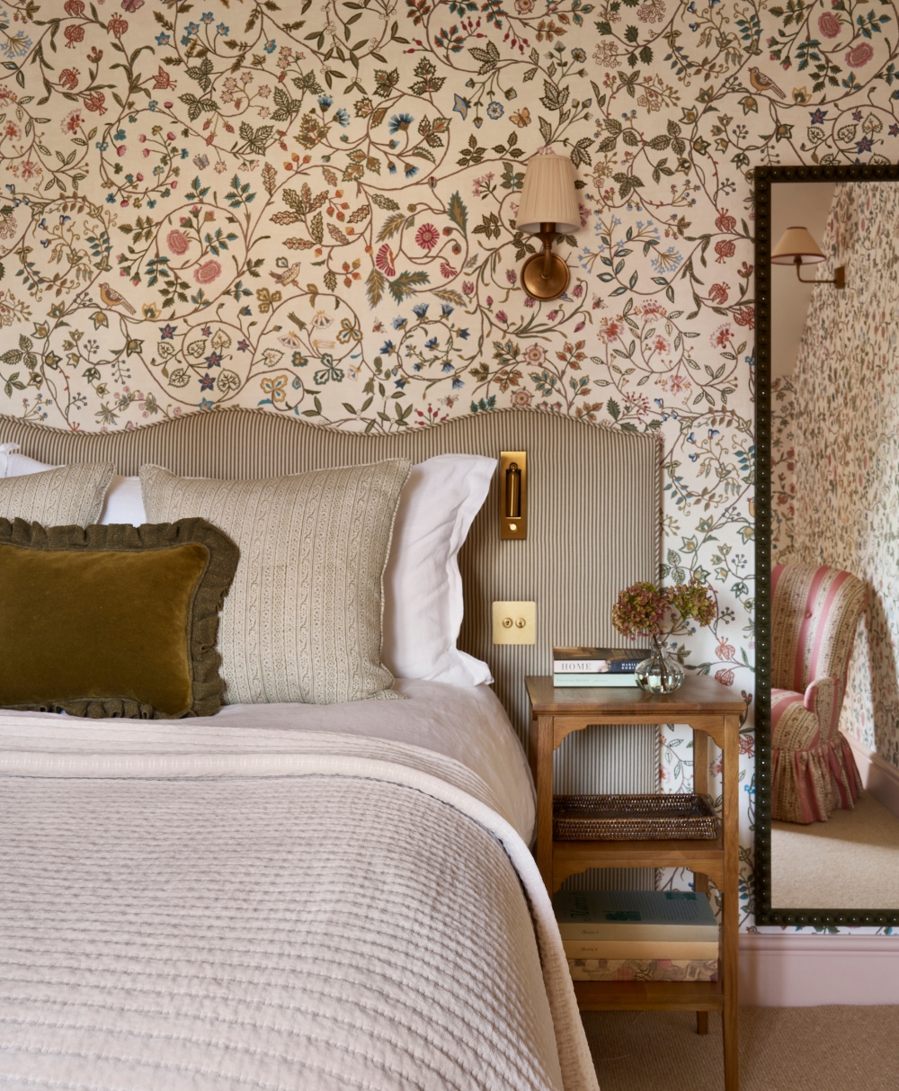 A cozy bedroom with a floral wallpaper, a bed with neutral bedding and a green accent pillow, a wooden bedside table with books and flowers, and a large mirror reflecting a pink-striped chair.