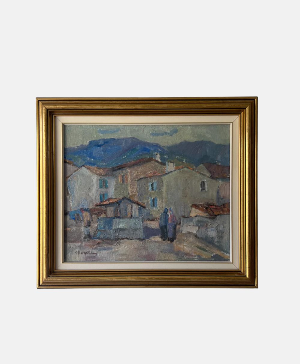 A framed painting depicting a village scene with houses, mountains in the background, and two figures walking in the foreground, painted in soft, muted colors.