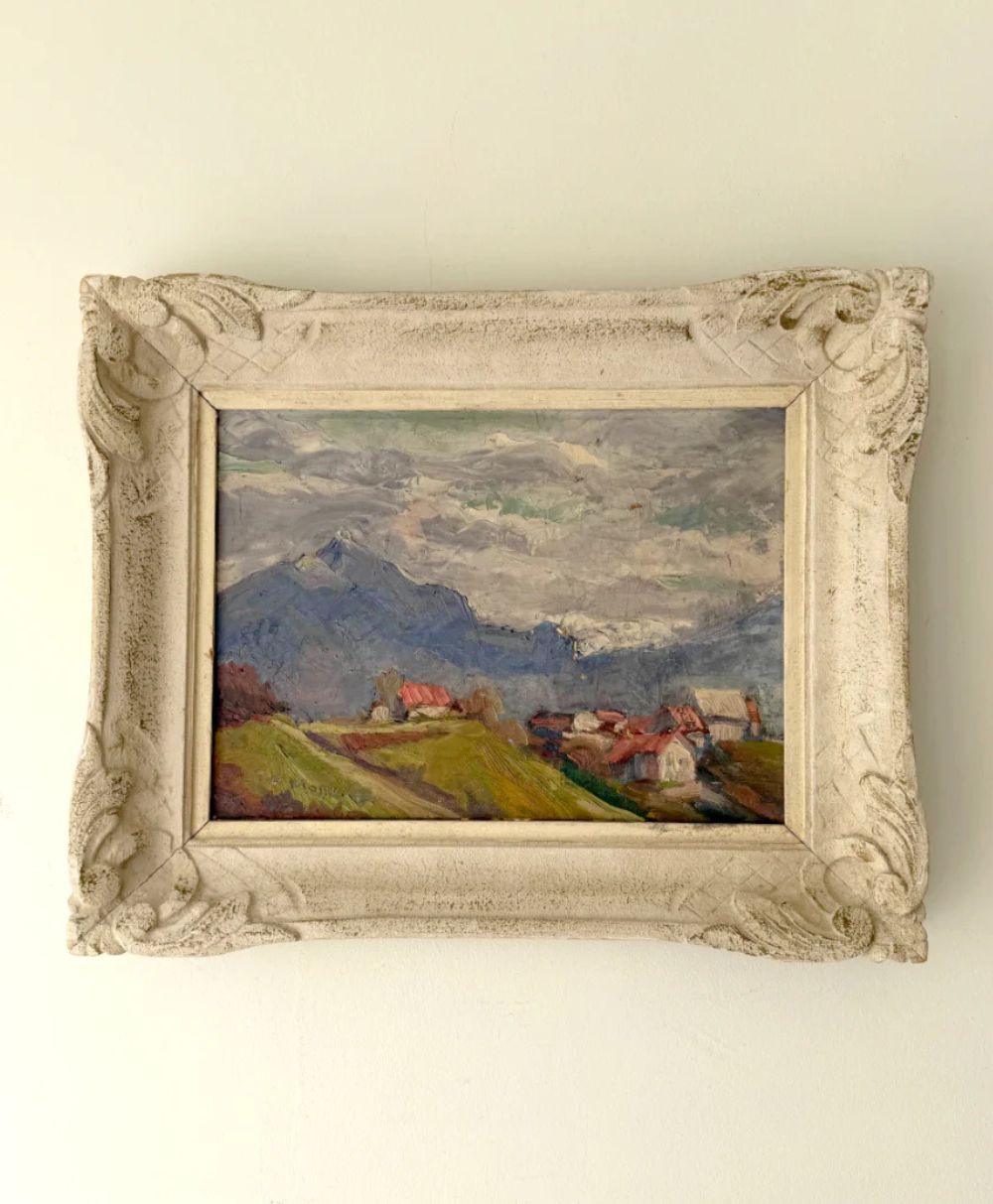 A framed landscape painting with mountains, cloudy sky, and a small village with red-roofed houses in the foreground, all in muted, earthy tones. The ornate frame is off-white and textured.