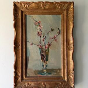 A framed painting of delicate pink flowers in a glass vase, done in an impressionist style with muted colors, hangs on a plain light-colored wall. The ornate wooden frame has decorative carvings.