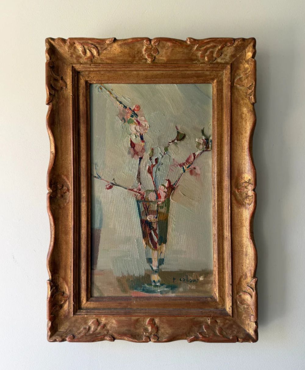 A framed painting of delicate pink flowers in a glass vase, done in an impressionist style with muted colors, hangs on a plain light-colored wall. The ornate wooden frame has decorative carvings.
