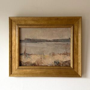 A framed landscape painting with earthy tones, depicting a calm body of water and dry grass under a cloudy sky, hangs on a plain white wall. The frame is gold and ornate.