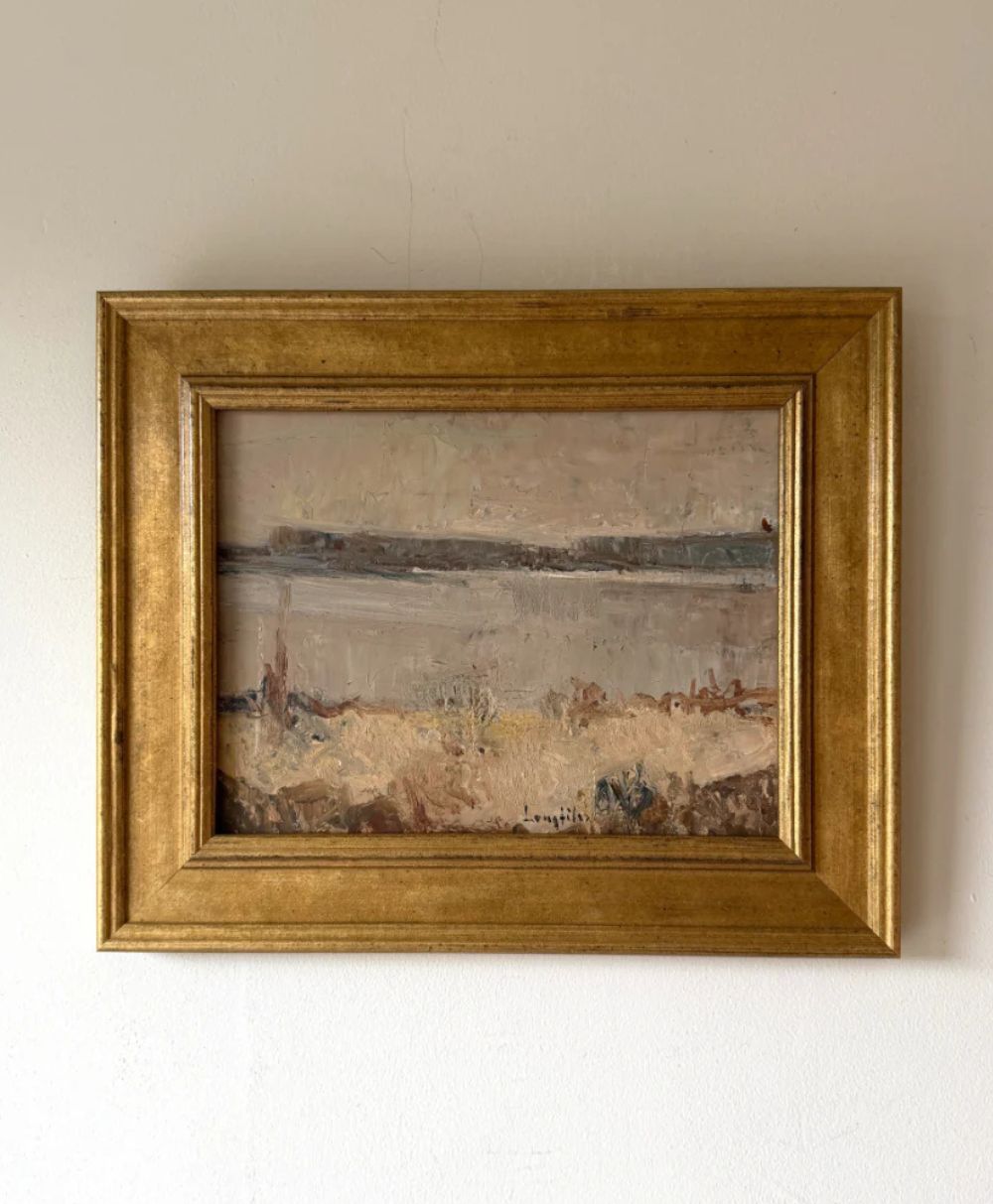 A framed landscape painting with earthy tones, depicting a calm body of water and dry grass under a cloudy sky, hangs on a plain white wall. The frame is gold and ornate.