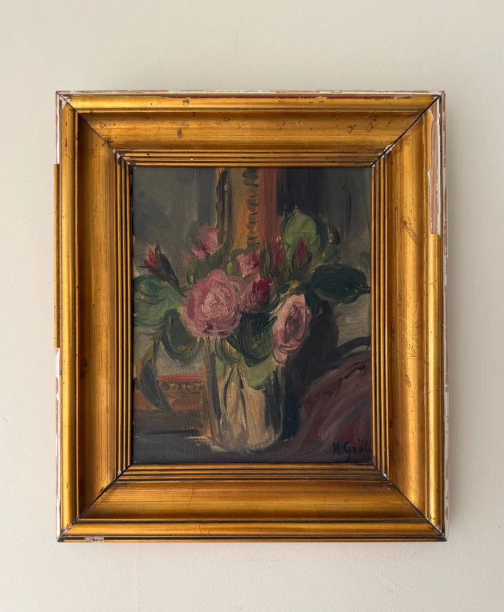 A framed painting of pink and red roses with green leaves in a vase, set against a dark background. The frame is gold with some visible wear and chipping along the edges.