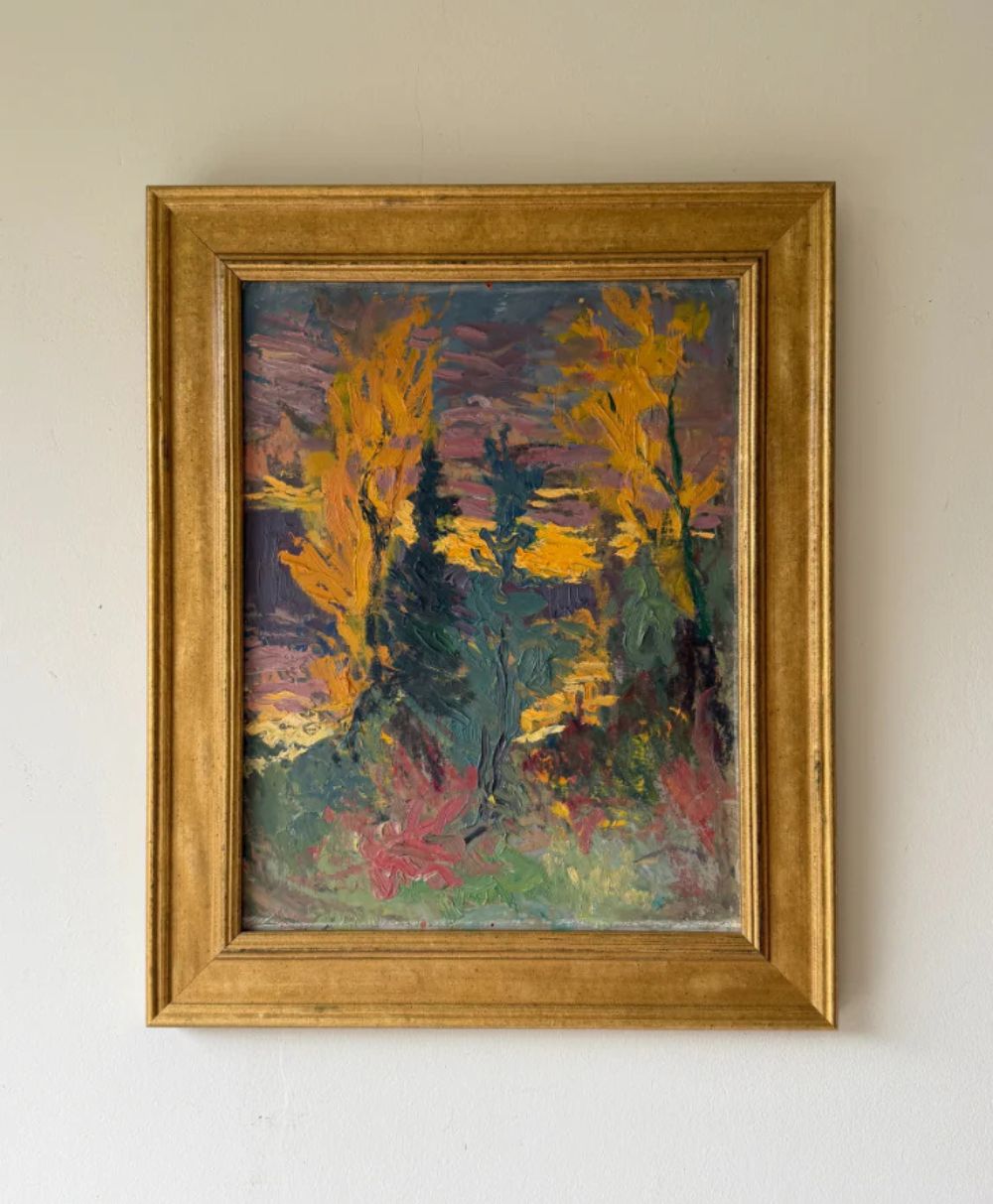Framed impressionist painting featuring bold, textured brushstrokes depicting yellow trees, green foliage, and a colorful sky in shades of purple, pink, and orange, hanging on a plain light-colored wall.