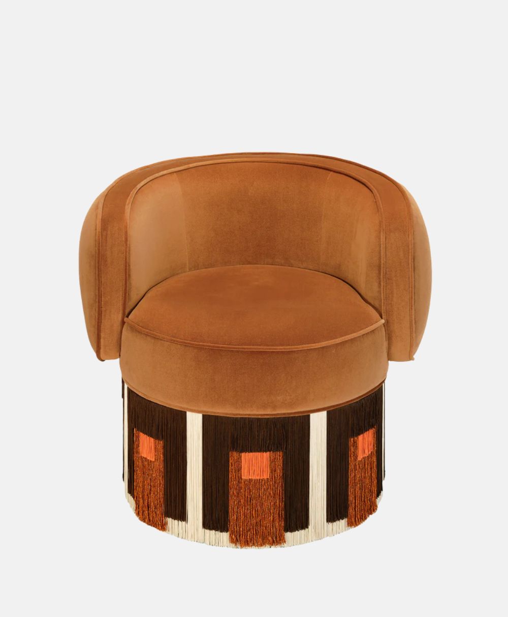 A modern, round, tan velvet chair with a curved backrest and colorful, geometric fringe detailing at the base in black, white, and orange. The background is plain white.