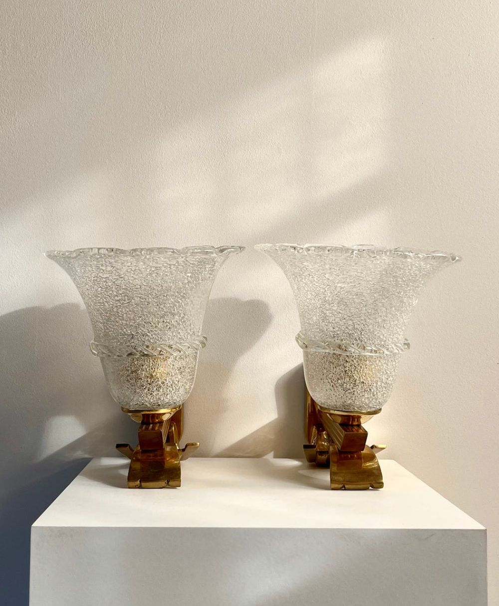 Two vintage glass wall sconces with textured, flared shades and brass bases are displayed side by side on a white pedestal against a neutral wall, illuminated by soft natural light.