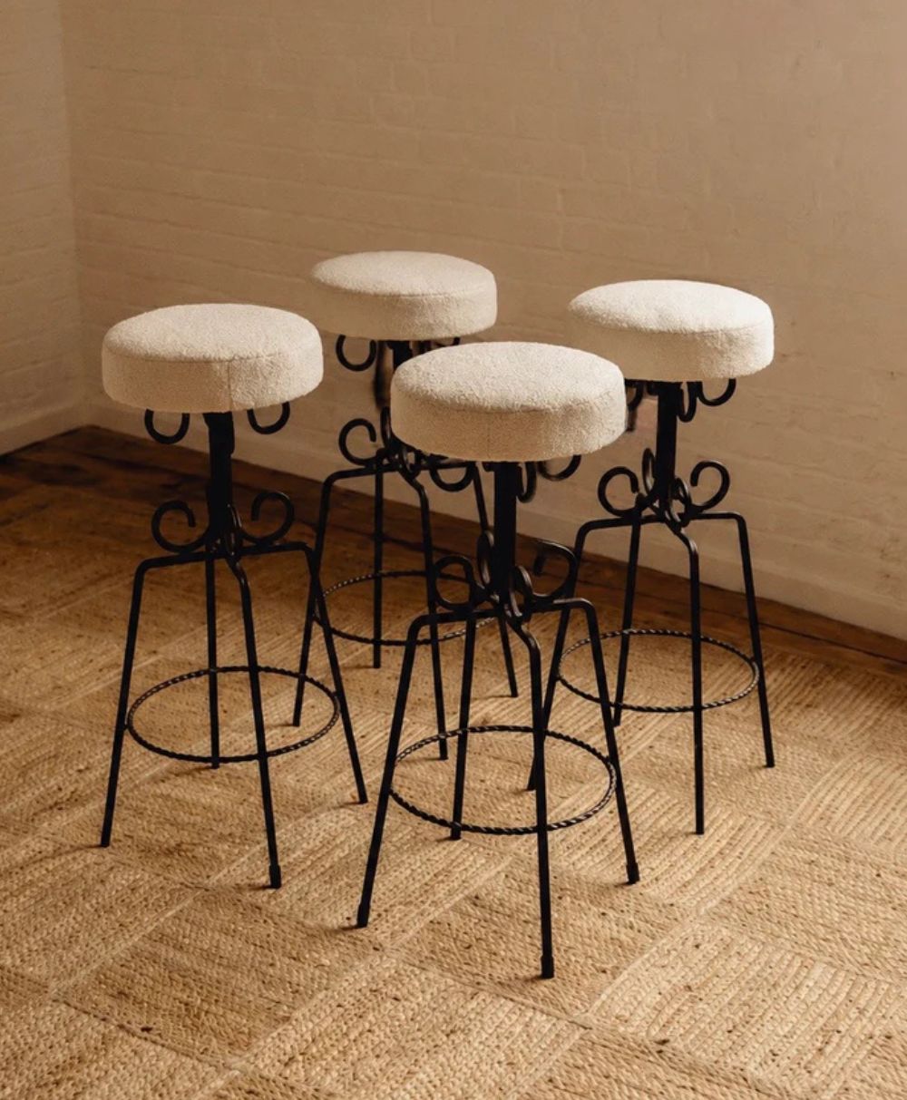 Four bar stools with metal frames and round, light-colored upholstered seats are arranged on a woven rug in a room with a white brick wall and wooden floor trim.
