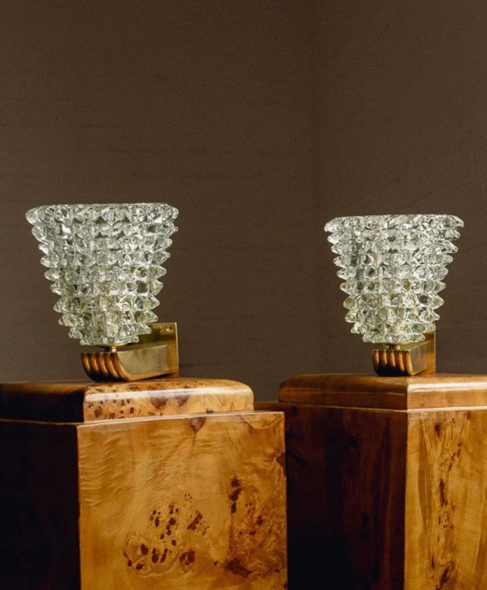 Two textured glass wall sconces with gold bases are mounted on wooden pedestals against a plain brown background. The glass shades have a geometric, diamond-like pattern and fan outwards from the brass fixtures.