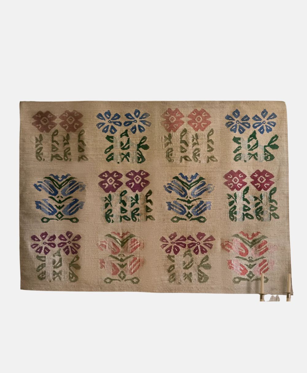 A woven textile featuring a grid of colorful, symmetrical floral patterns in red, blue, green, and purple on a beige background. The textile has a simple, folk-art style with repeating flower motifs.