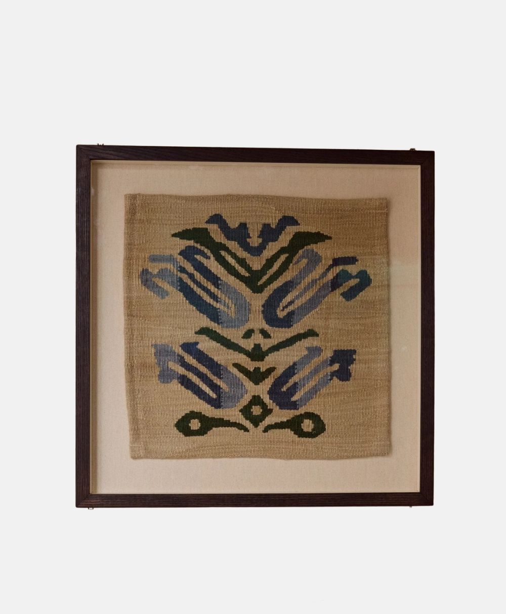 A framed textile artwork featuring a symmetrical, abstract pattern in blue, green, and dark tones on a beige background, displayed against a light wall.