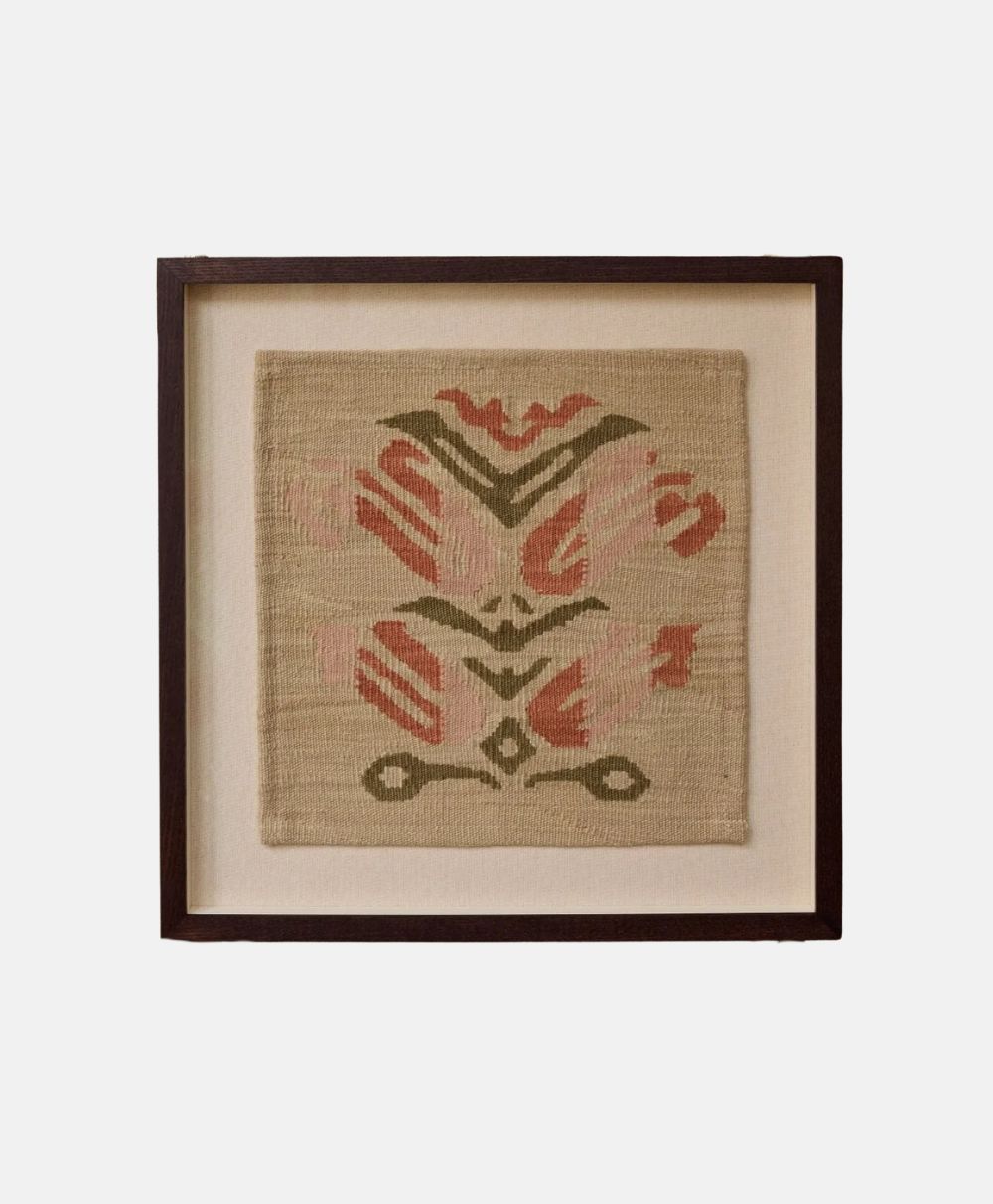 A square piece of woven beige fabric with a geometric pattern in red and green, displayed in a dark wooden frame against a light background.