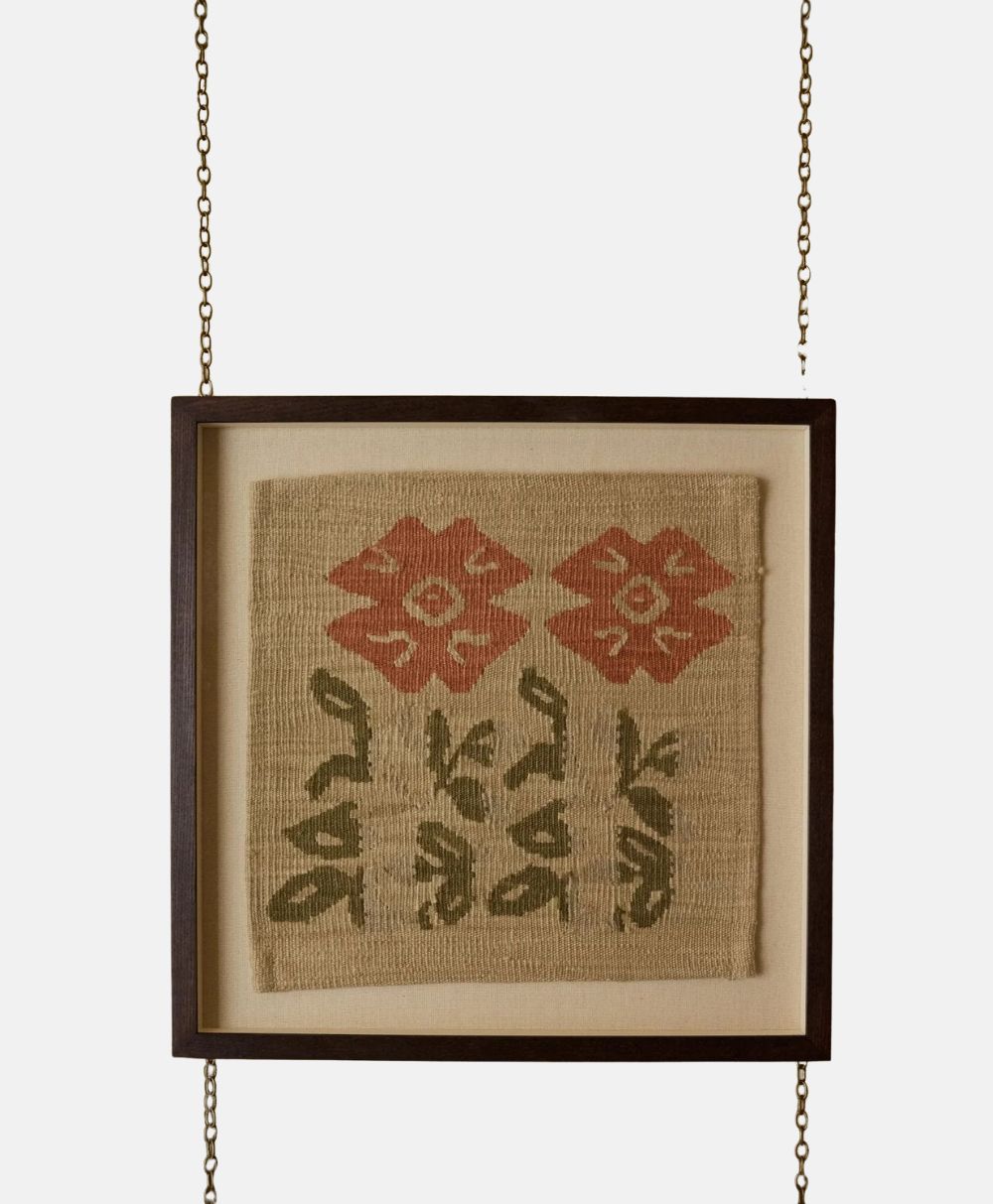 A framed textile art piece with two red floral patterns above green leafy stems, displayed in a dark wooden frame suspended by two metal chains.