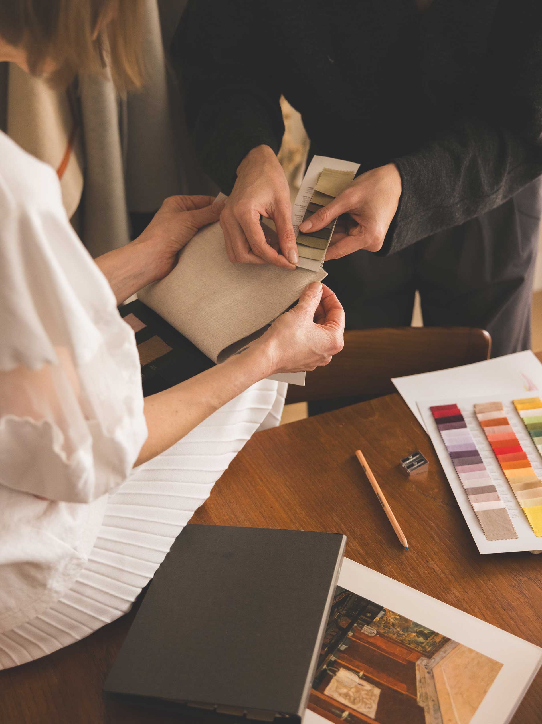 Two people examine a fabric swatch together at a table with a color palette, pencil, and design materials spread out, suggesting a collaborative design or interior decorating process.