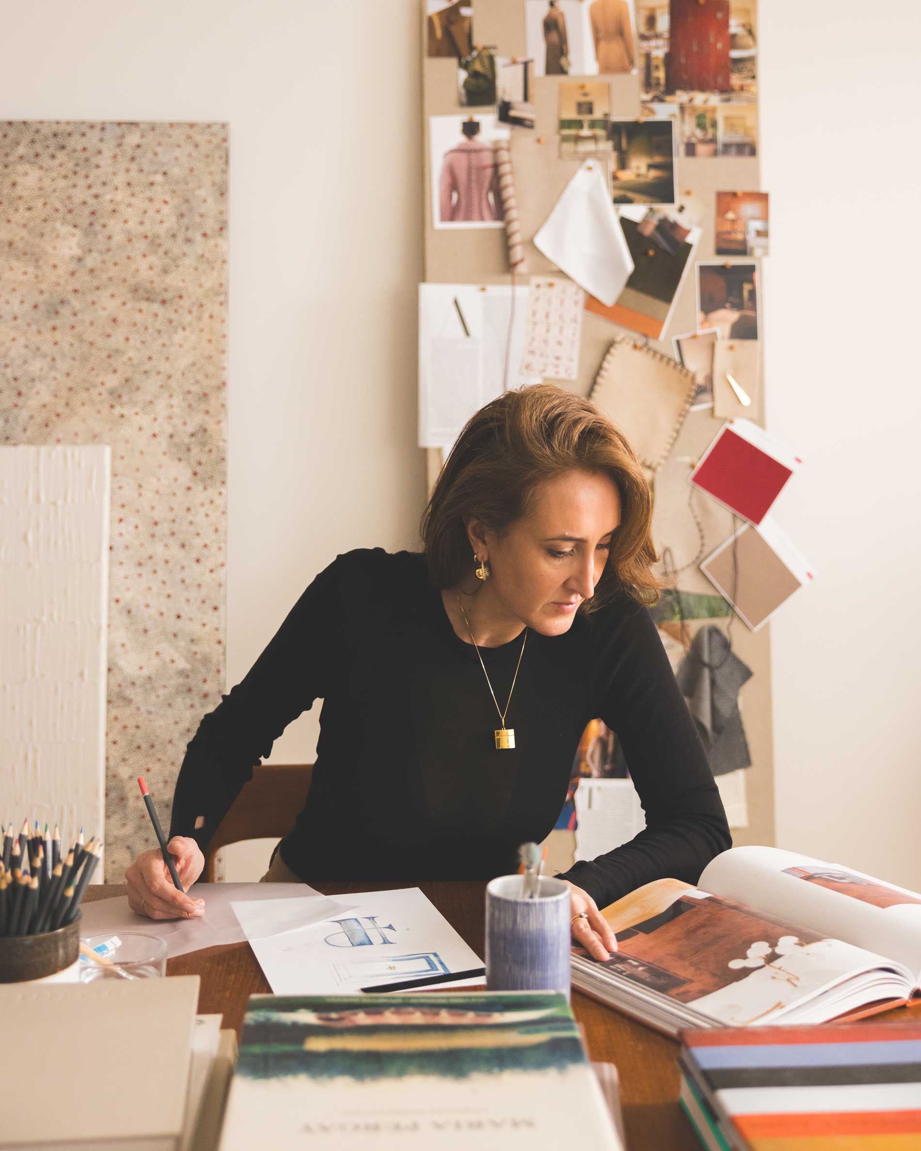 A woman with long brown hair sits at a desk, sketching on paper. She is surrounded by books, a candle, and art supplies, with a creative mood board full of images, fabric swatches, and notes behind her.