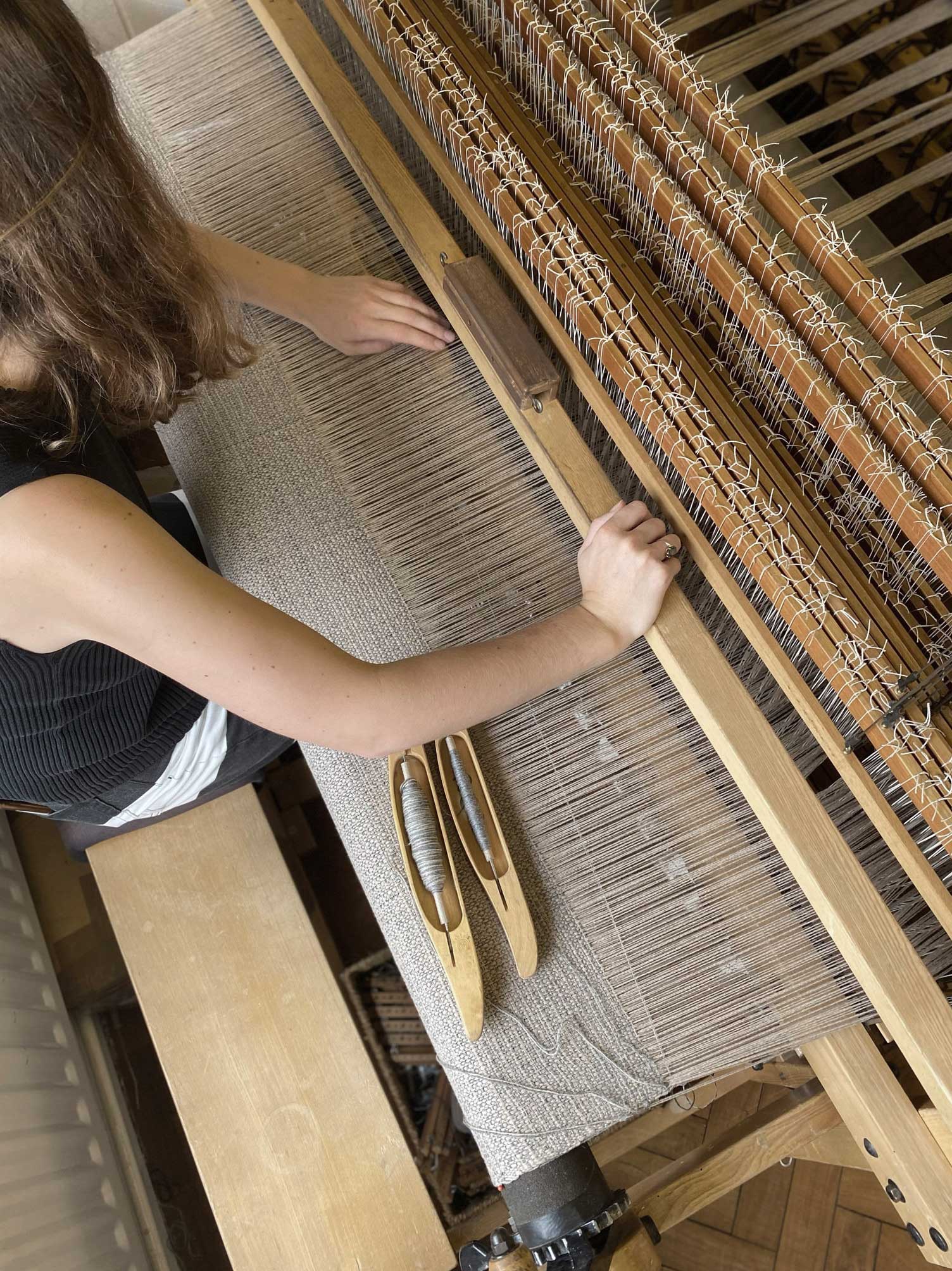 A person with long brown hair is weaving fabric on a wooden loom, using both hands to guide the threads. Two wooden shuttles are resting on the partially woven textile.