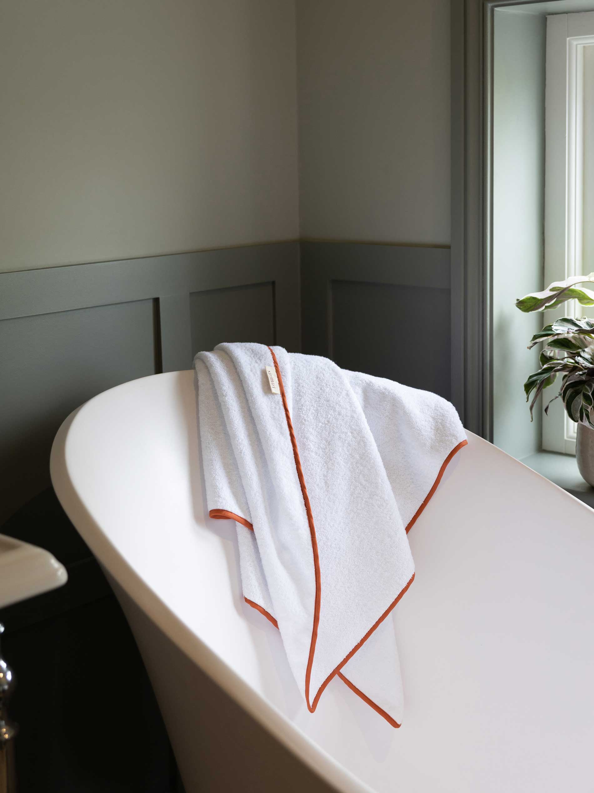 A white towel with orange trim is draped over the edge of a white freestanding bathtub near a window with light streaming in. A green potted plant sits on the windowsill.
