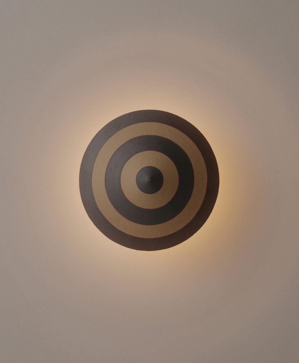 A round wall sconce with concentric black and brown circles emits a soft, warm glow onto a light-colored wall, creating a halo effect around the fixture.