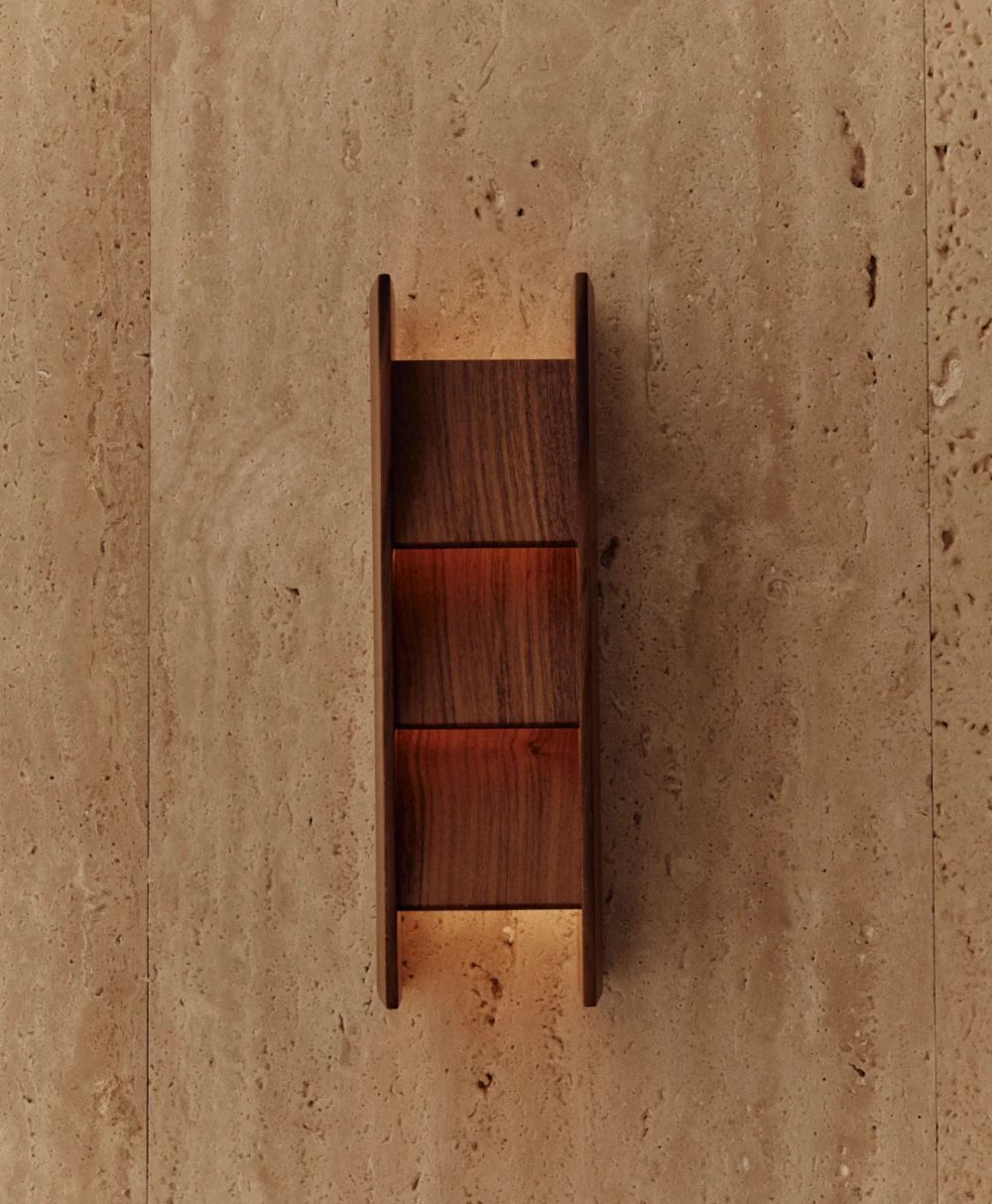 A modern wooden wall sconce with vertical panels emits a warm light, mounted on a beige stone wall with a textured, natural finish.